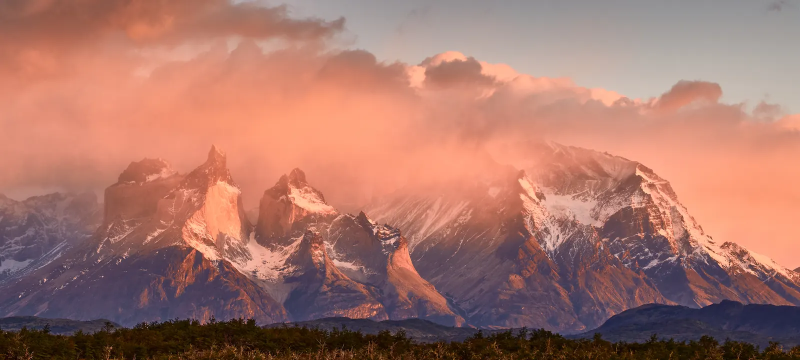 Coucher de soleil sur les Cuernos del Paine : les parois sombres et les sommets enneigés du massif du Paine sous des nuages orangés, au-dessus d’un plan d’eau.